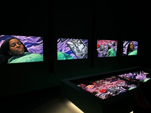 Four bright photographs projected in a dark room showing a Black woman lying with eyes closed on purple fabric