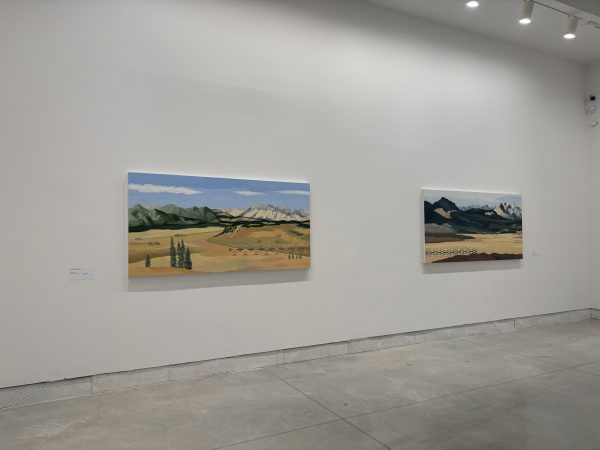 Two paintings hang on white gallery walls: desert and mountain scenes