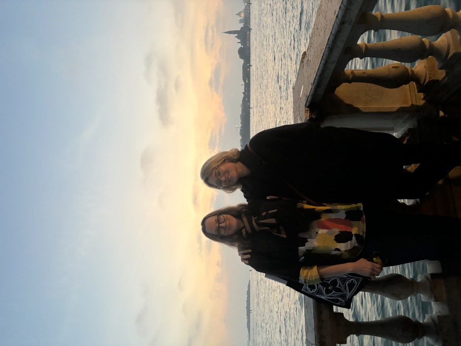 Two women stand in front of a rippling body of water and silhouette of Venice as sunset falls.