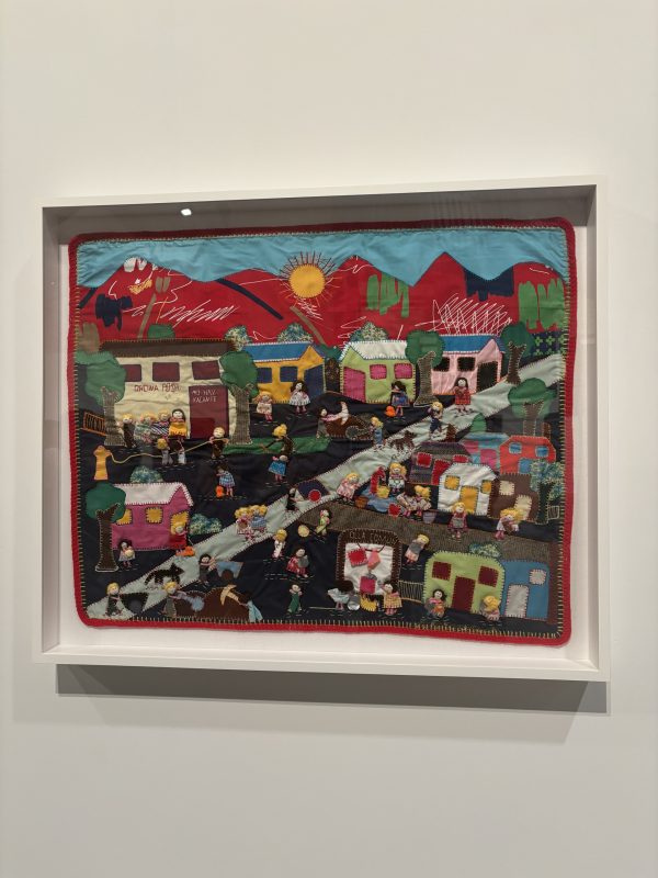 A framed hand-sewn textile artwork with block colours and visible stitches, depicting a community playing and working on the streets against a backdrop of mountains and blue sky