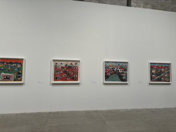 A display of four hand-sewn textile artworks hang on a white gallery wall. They depict community scenes; people on streets, outside their houses, playing and working. A church is visible in one; a pond with sailing boats in another; a dove with a banner in another.