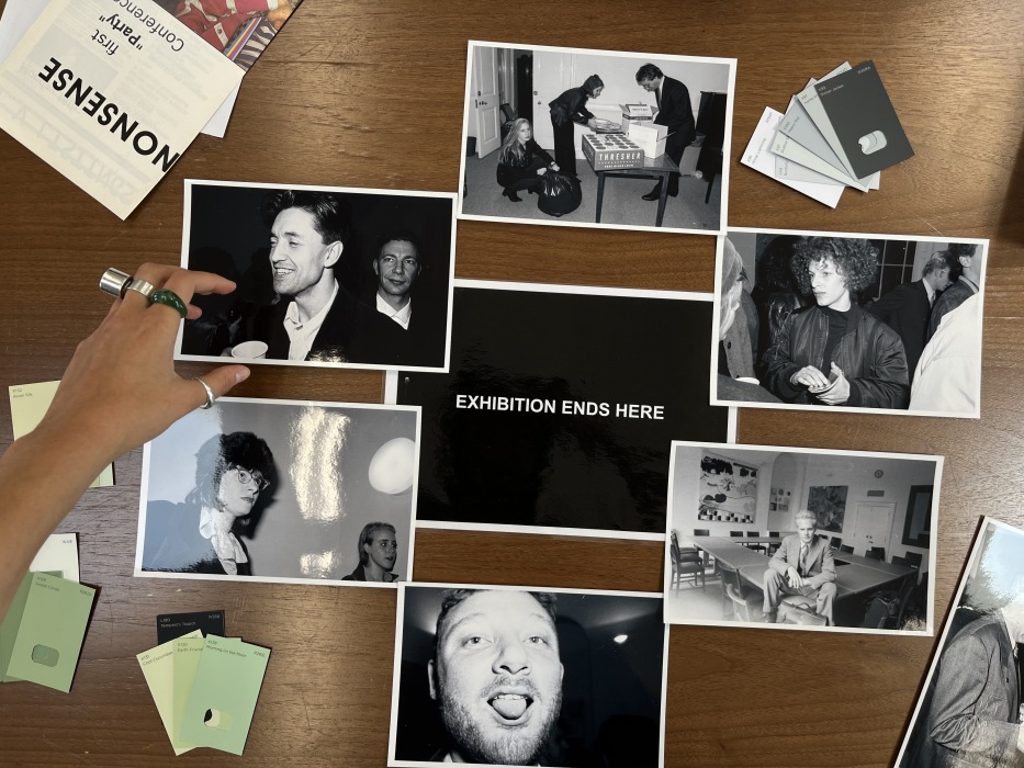 An image taken from above of black and white photographs spread out on a table; a hand arranges them. The centre photograph reads 'EXHIBITION ENDS HERE'.