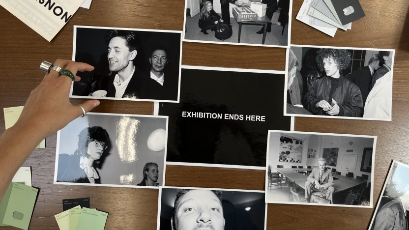 An image taken from above of black and white photographs spread out on a table; a hand arranges them. The centre photograph reads 'EXHIBITION ENDS HERE'.