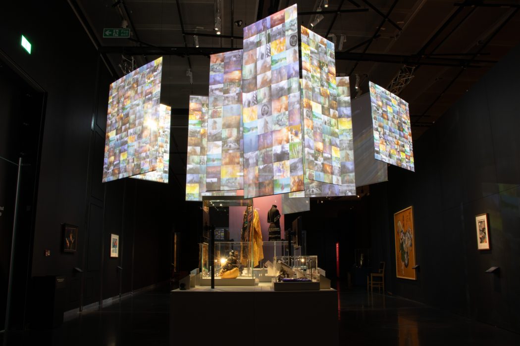 Installation view of Samurai Exhibition at the British Museum, featuring suspended screens displaying a patchwork of images above a central display of objects and garments.