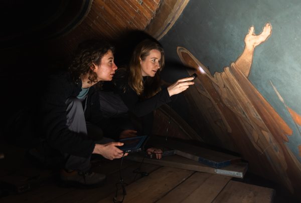 Two women crouch beside a wall painting. One holds a small torch to the surface. The painting is of a figure with arms spread.