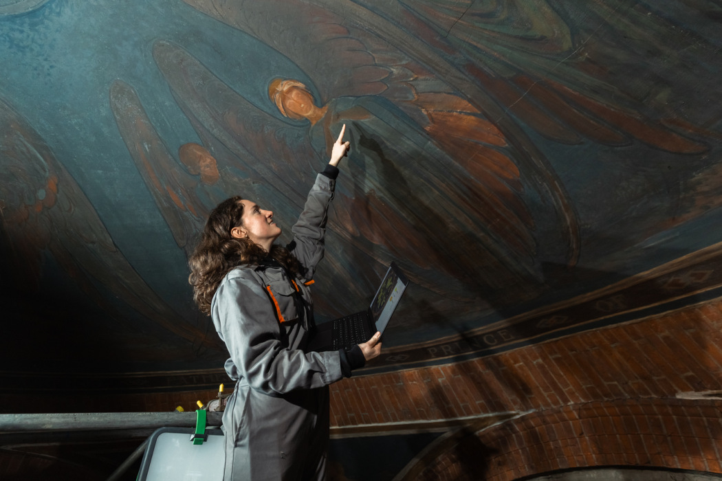 A woman in a jumpsuit points at paintings on a domed ceiling, painted with blue and orange winged women. She has a laptop in one hand and is tied to scaffolding just visible in the corner.