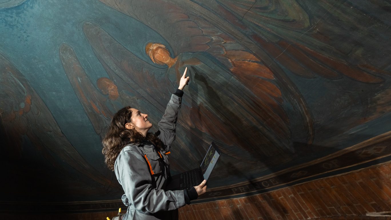 A woman in a jumpsuit points at paintings on a domed ceiling, painted with blue and orange winged women. She has a laptop in one hand and is tied to scaffolding just visible in the corner.