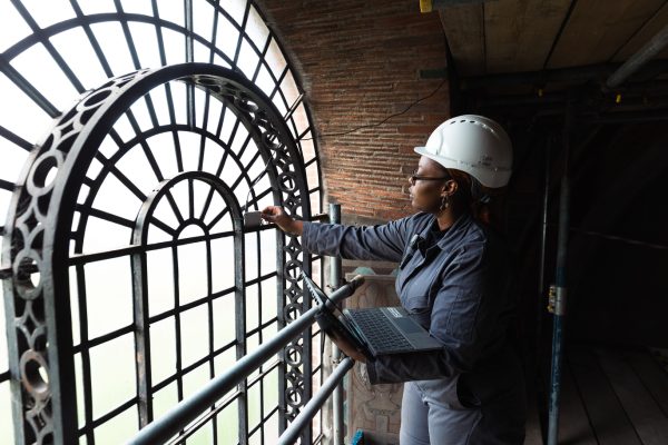 A student in a hard hat and jumpsuit holds a laptop. She stands beside a bright arched window. She is placing a monitor on the window.