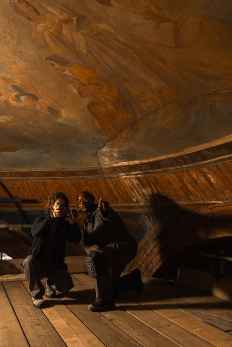 Two women crouch on bare planks, with monitoring equipment facing the camera. Above their heads, a dome of wall paintings.