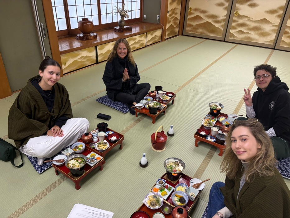 Four people sit on mats on the floor with meals in front of them on wooden trays, and a teapot between them.