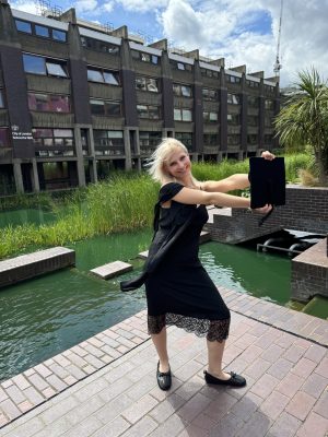 A woman stands in front of a Brutalist building, water and plants. She has bleach blonde hair and a big smile, and she's wearing a graduation gown. She holds her cap out in front of her.