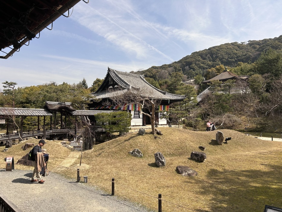 A Japanese building sits in front of a mountain covered in forest. Some flags are strung around the building. It is a sunny day.