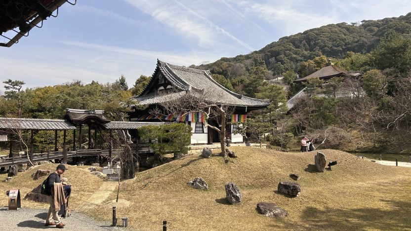 A Japanese building sits in front of a mountain covered in forest. Some flags are strung around the building. It is a sunny day.