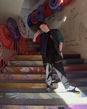A man stands on a staircase doing a peace sign. The staircase is brightly patterned and a display of parasols are hung behind him.