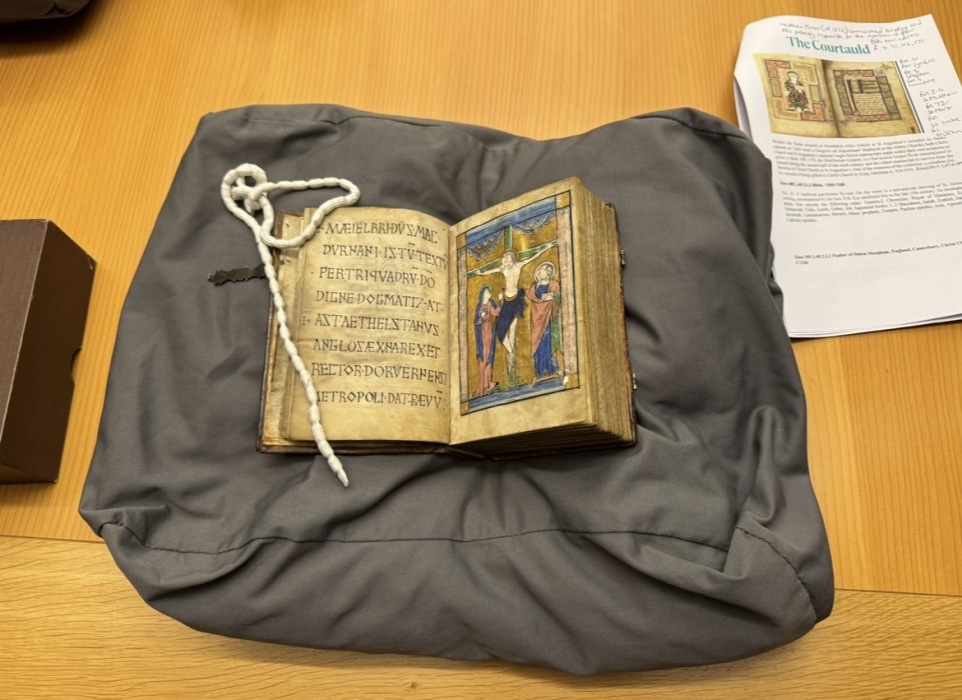 A small book rests on black cloth on a table. The book is open to a page of medieval writing, and a small, golden image of Christ on the cross. Next to the book is a worksheet from the Courtauld Institute.