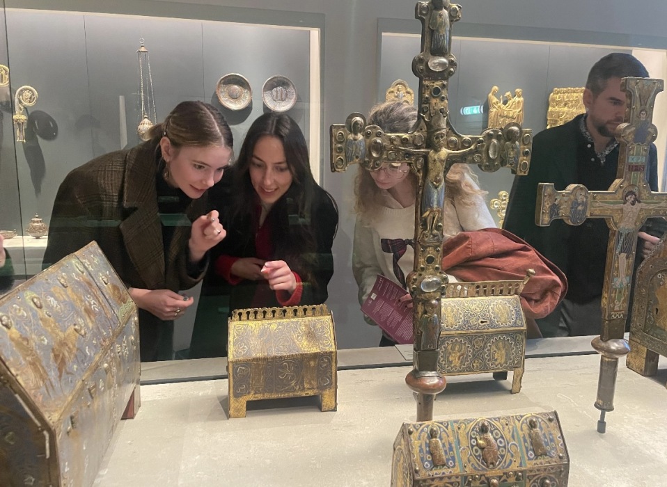 Students peer at enamel relic boxes and depictions of Christ on the cross in a display case, seeming to discuss the objects.