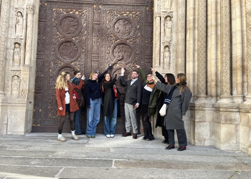 A group of smiling students stand outside an ornate door, pointing at a particular relief on it