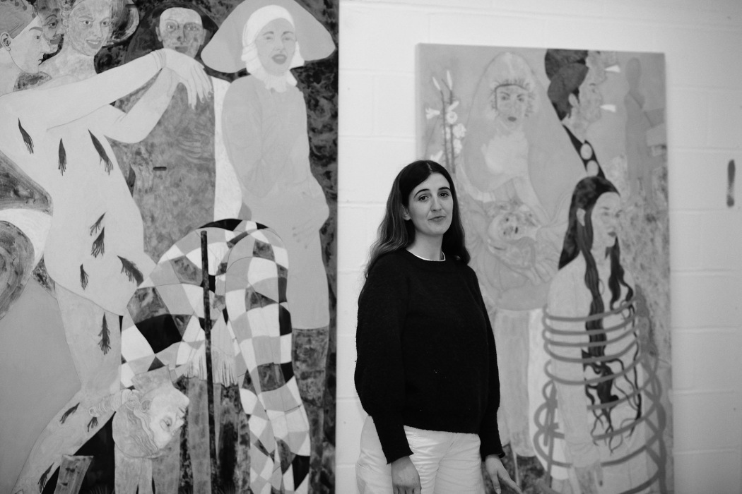 A black and white portrait of artist Ella Walker standing in front of canvases