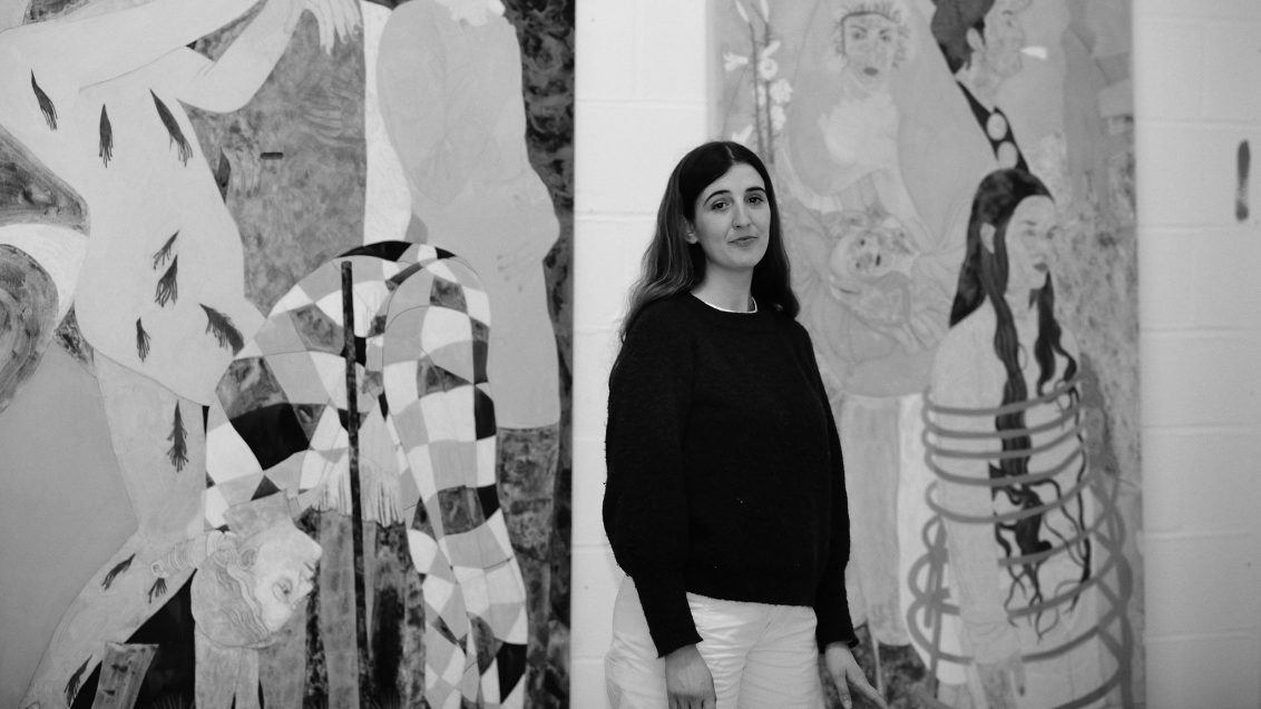 A black and white portrait of artist Ella Walker standing in front of canvases