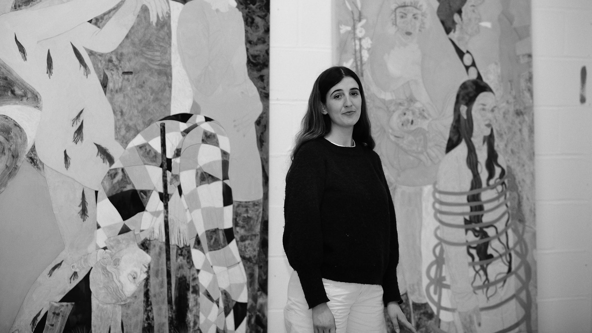 A black and white portrait of artist Ella Walker standing in front of canvases