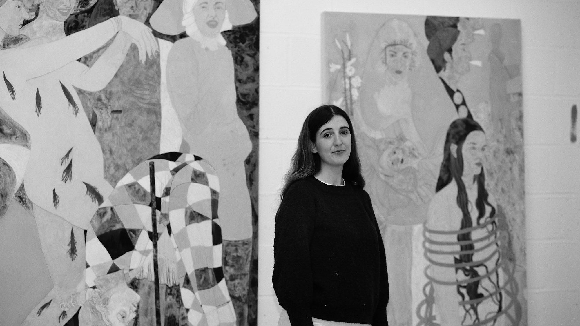 A black and white portrait of artist Ella Walker standing in front of canvases