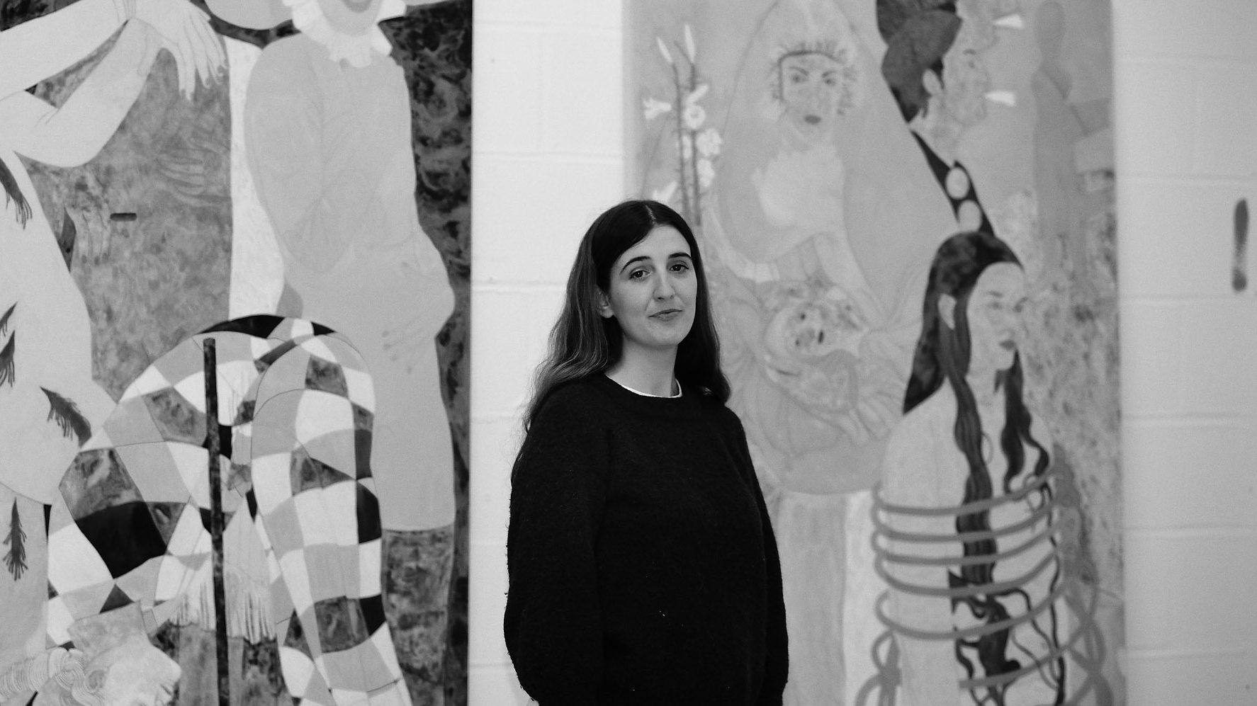 A black and white portrait of artist Ella Walker standing in front of canvases
