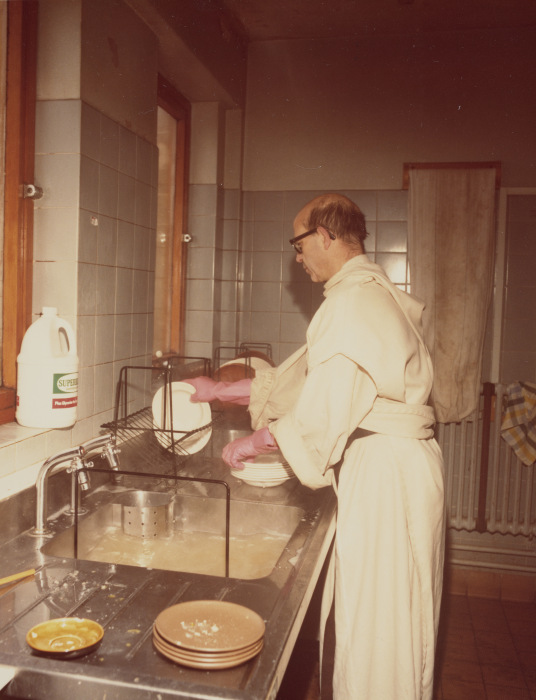 A sepia photograph of a man in a white dressing gown and pink rubber gloves. There are dirty dishes in the foreground - he is doing the washing up.