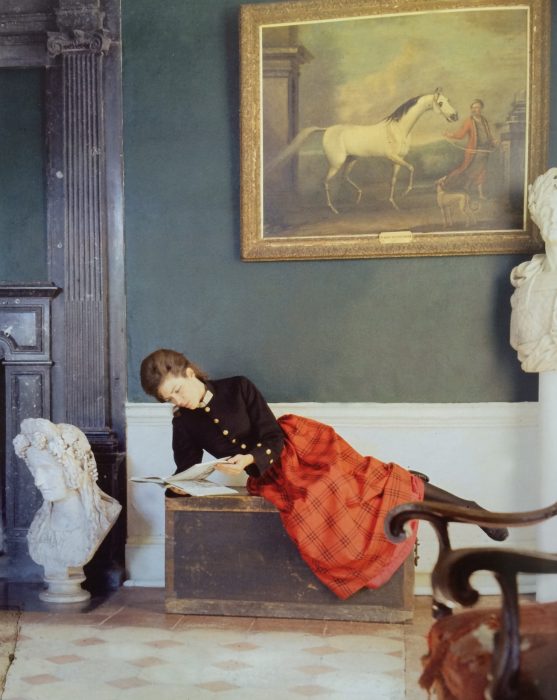 a woman reclines in an ornate interior reading. She wears a bright ornate tartan skirt and black formal top.