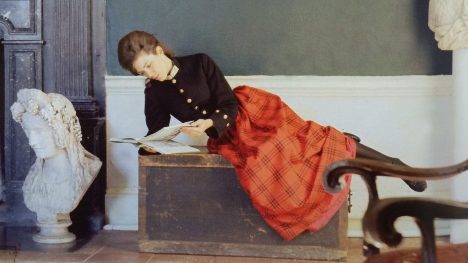 a woman reclines in an ornate interior reading. She wears a bright ornate tartan skirt and black formal top.
