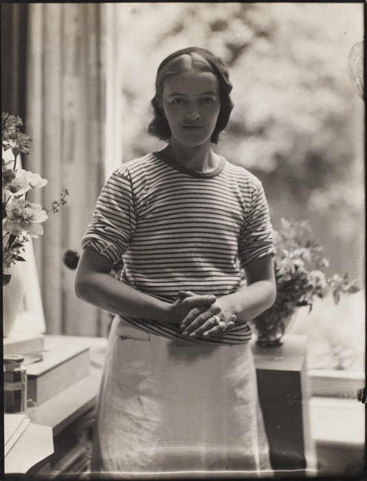 Black and white photograph of Barbara Hepworth in her studio. She wears a white skirt and a striped short sleeve top, with her hands clasped in front of her.