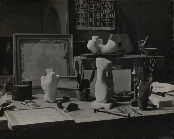 Black and white photograph of the studio, with three small white sculptures, an easel with a fabric work, and various tools dotted around a table.