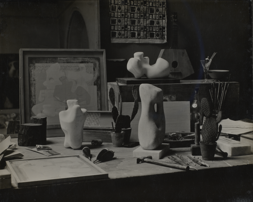 Black and white photograph of the studio, with three small white sculptures, an easel with a fabric work, and various tools dotted around a table.