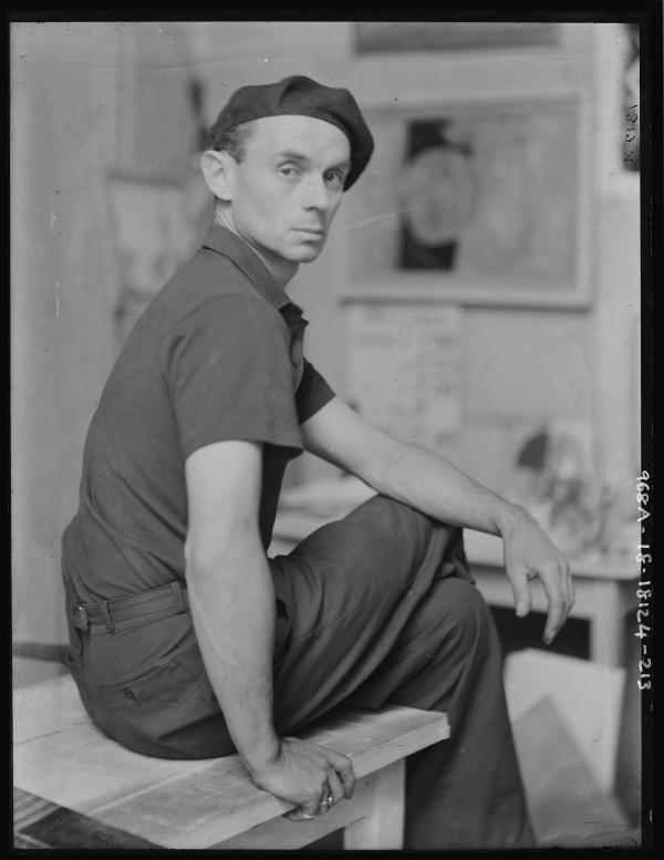 Black and white photograph of Ben Nicholson. He wears dark trousers and top with a flat cap. He sits posed, with legs crossed, looking away from the camera on his studio table.