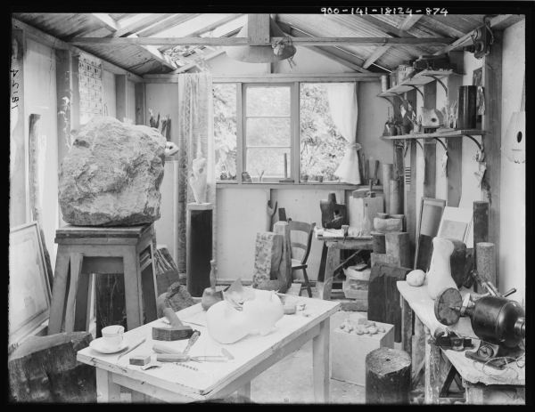 Black and white photograph of a busy art studio, with a carving table with tools in front of lots of sculptures visible behind. The studio is light filled, with skylights and a large window at the end.