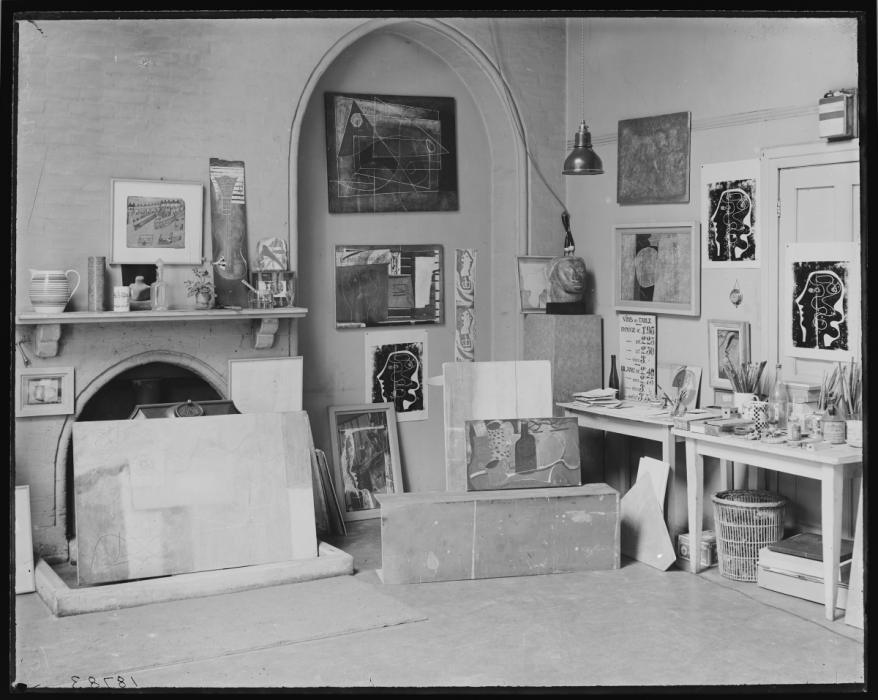 Black and white photograph of an indoor studio, fulled with artworks, sculptures, tools, plants, and glassware.