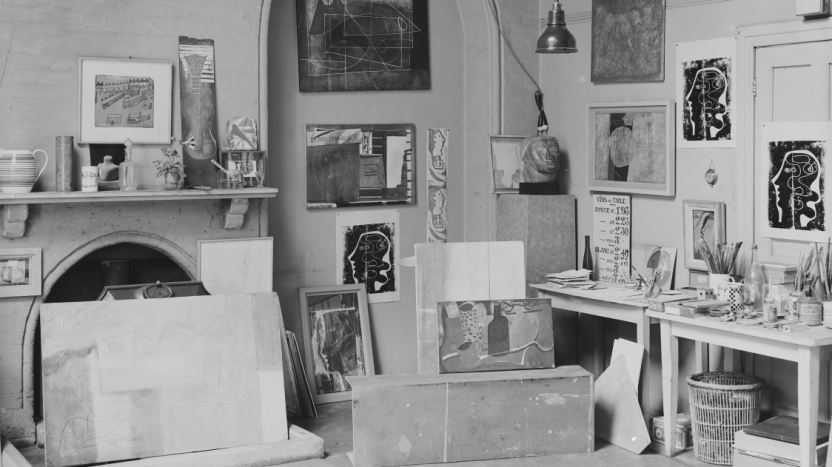 Black and white photograph of an indoor studio, fulled with artworks, sculptures, tools, plants, and glassware.