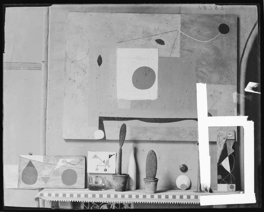 Black and white photograph of a mantlepiece with an abstract artwork hung above, and many small carvings below. One is circled on screen; Nicholson's first relief.