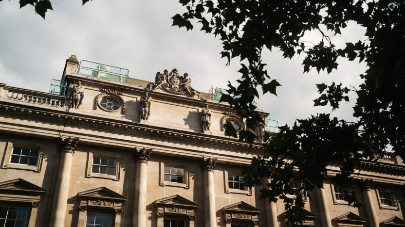 Exterior view of the historic Somerset House stone building with ornate architectural details and statues, partially framed by tree branches.