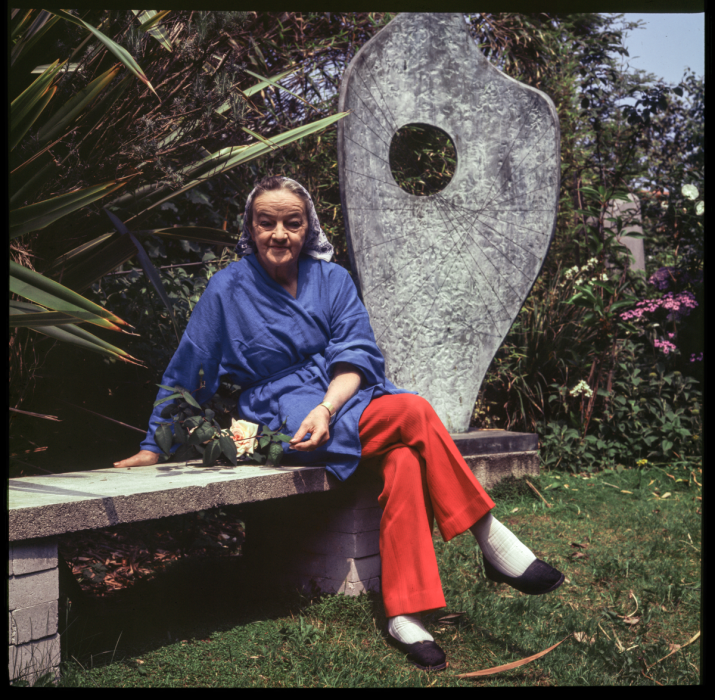 Photograph of Barbara Hepworth wearing a blue jacket and bright red trousers. She is sitting on a bench in a garden, with a large curved sculpture behind her.