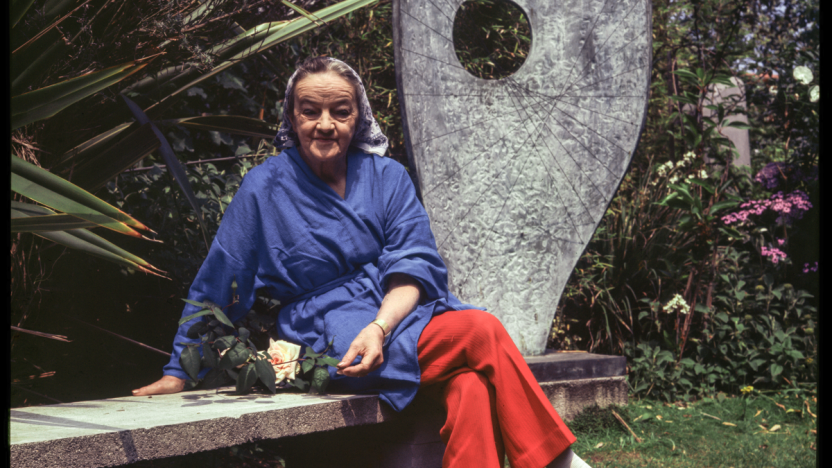 Photograph of Barbara Hepworth wearing a blue jacket and bright red trousers. She is sitting on a bench in a garden, with a large curved sculpture behind her.
