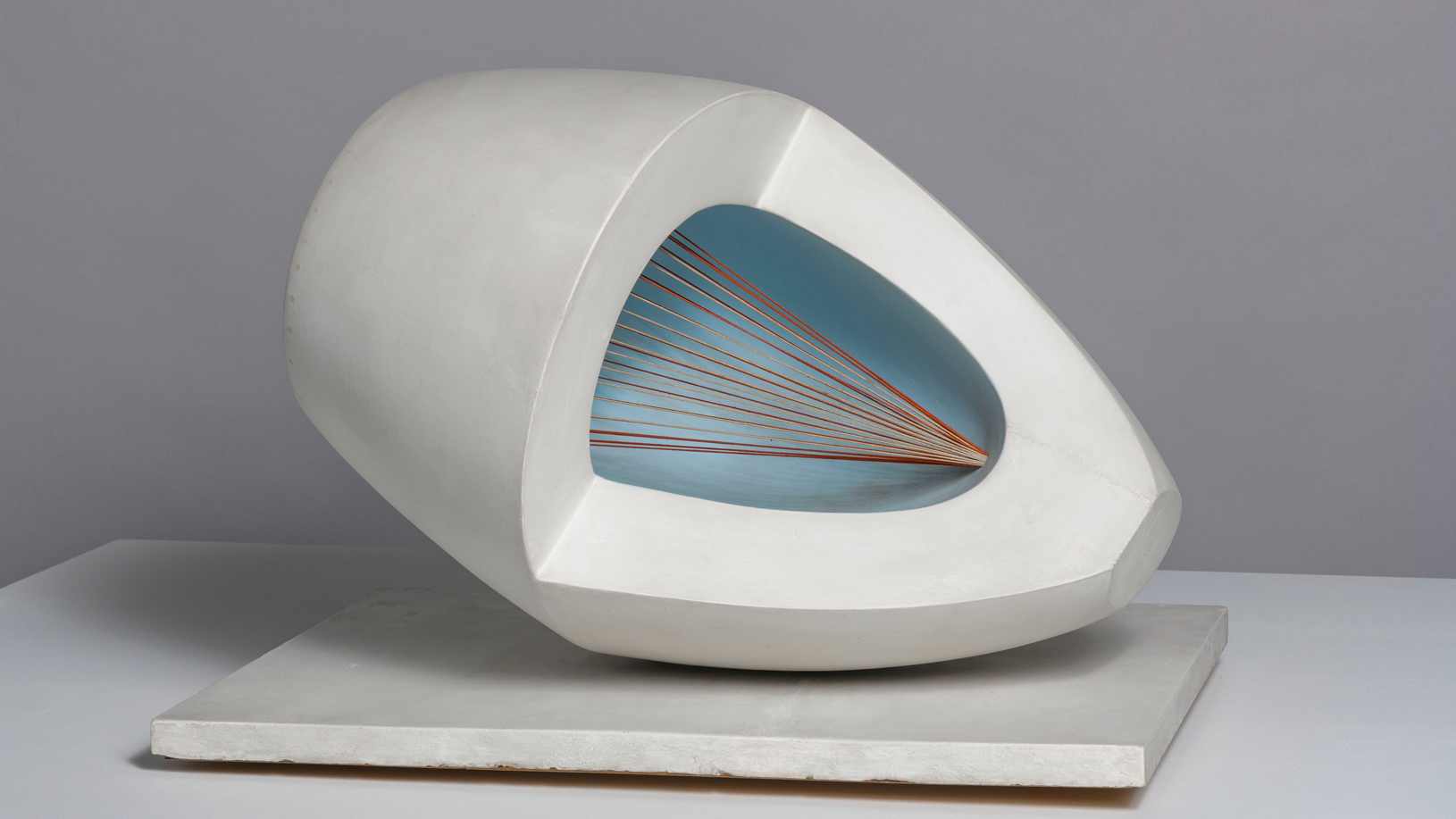 a wooden abstract sculpture featuring an oval form with pale blue and red painted surfaces, intersected by strings and mounted on a painted wooden base.
