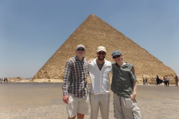Three men in hats and sunglasses stand in front of a pyramid in Egypt.