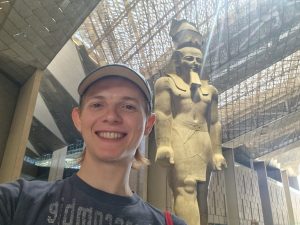 A man smiles in front of an Egyptian statue in a museum