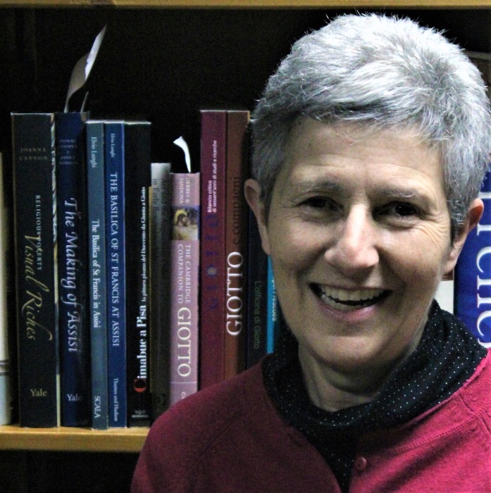 Smiling woman with short white hair wearing a red shirt stands in front of a bookshelf.