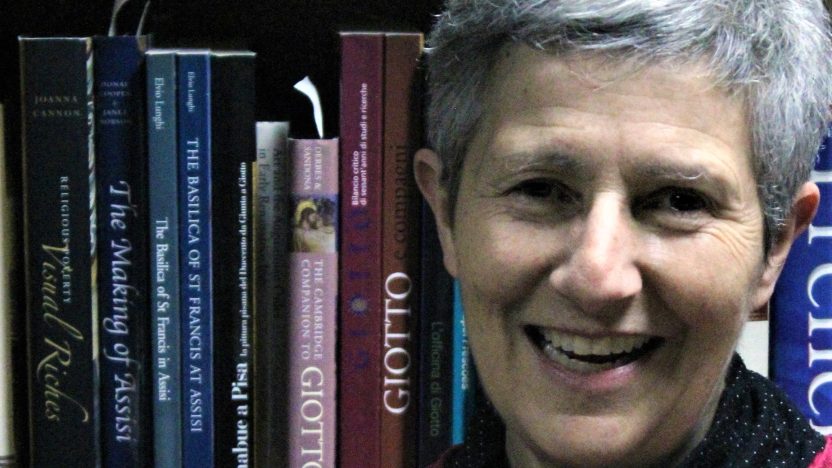 Smiling woman with short white hair wearing a red shirt stands in front of a bookshelf.