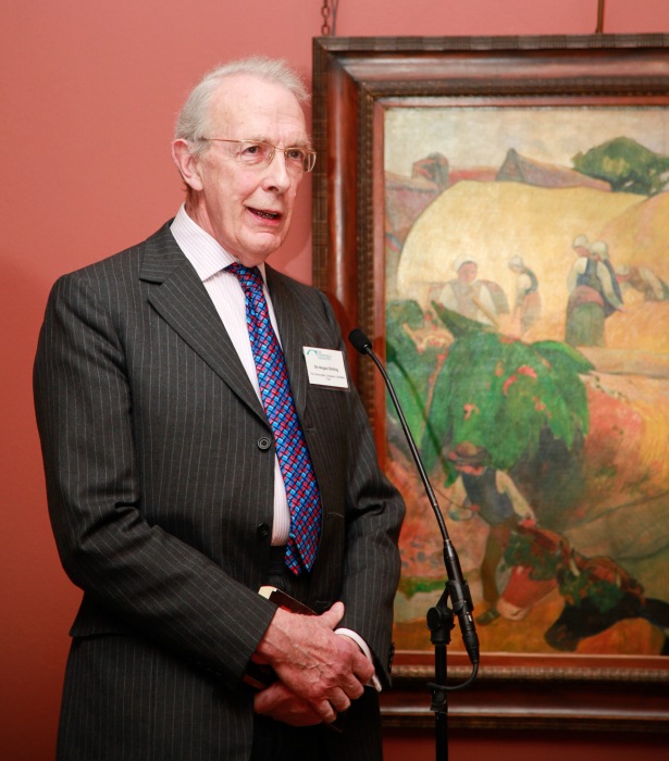 Angus Stirling standing in front of a framed Gauguin painting on a salmon-colour painted wall.