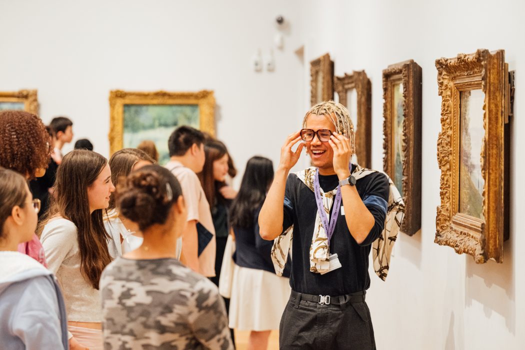 A teacher is stood by an artwork, speaking to a group of teenage school students.