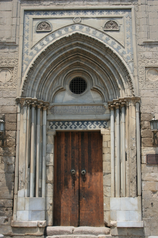 An ornate doorway. The doors are wooden, above them is elaborate white stone carving including pillars and a pattern in black and white.