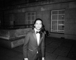 Black and white film photograph of a man in a suit, looking towards the camera, moving through a neoclassical looking courtyard