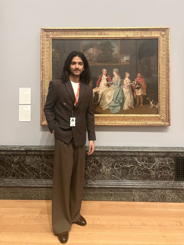 A man with long dark hair wearing a red lanyard stands in front of an 18th century painting